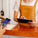 QR Codes - Crop anonymous female employee with application on cellphone screen interacting with partner using tablet at counter in cafeteria